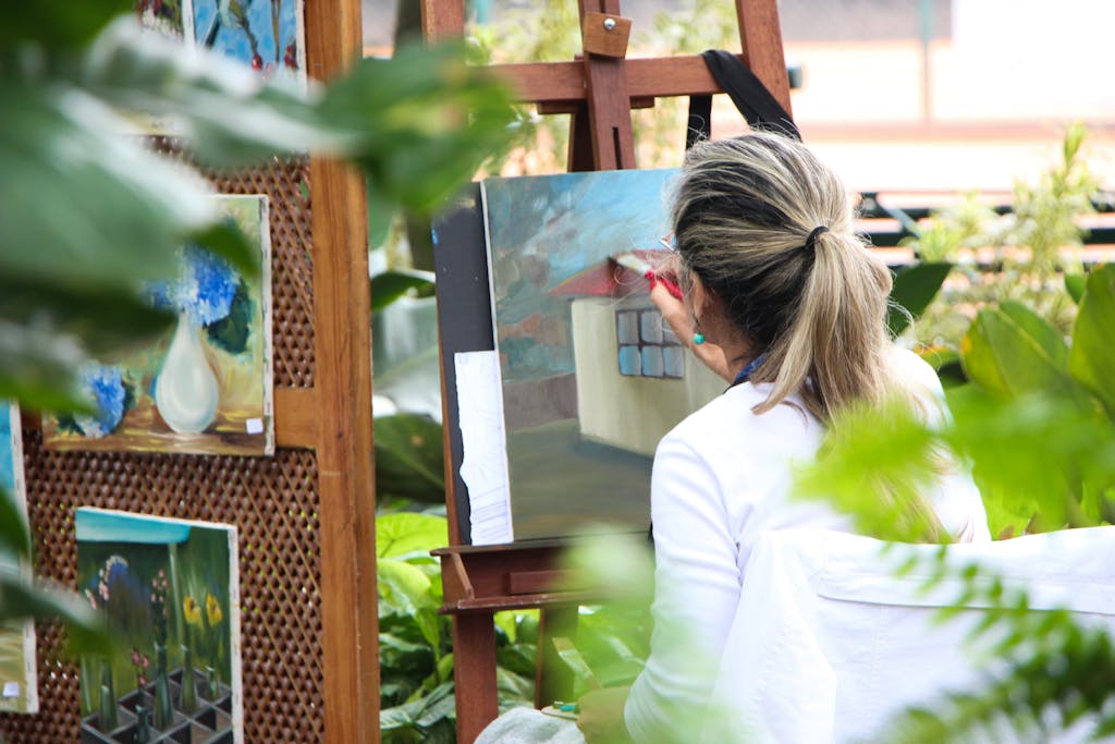 A woman enjoys painting in a lush outdoor garden, surrounded by vibrant artwork.