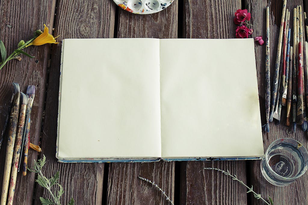 Blank sketchbook surrounded by paintbrushes and flowers on rustic wooden surface.