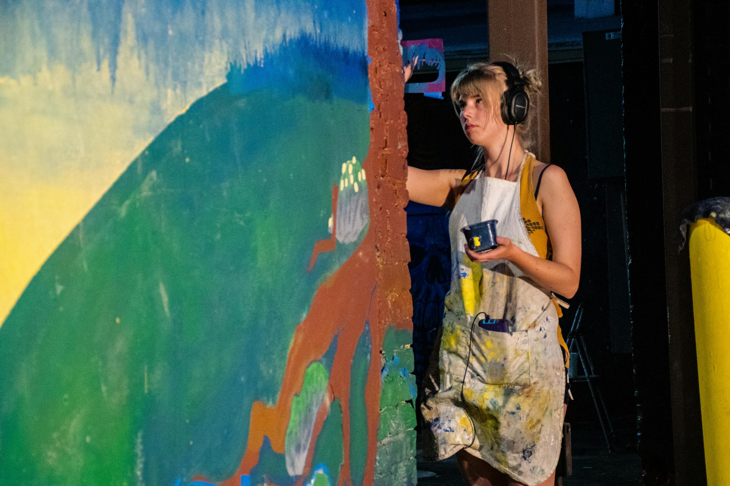 Female artist painting a vibrant mural while listening to music in an indoor studio environment.